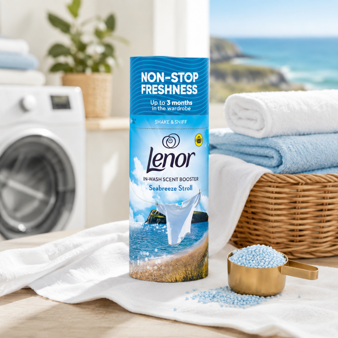 A bottle of Lenor Non-Stop Freshness Sea breeze Stroll in-wash scent booster stands on a white towel in a bright laundry room setting with a washing machine, folded towels, and blue detergent beads.