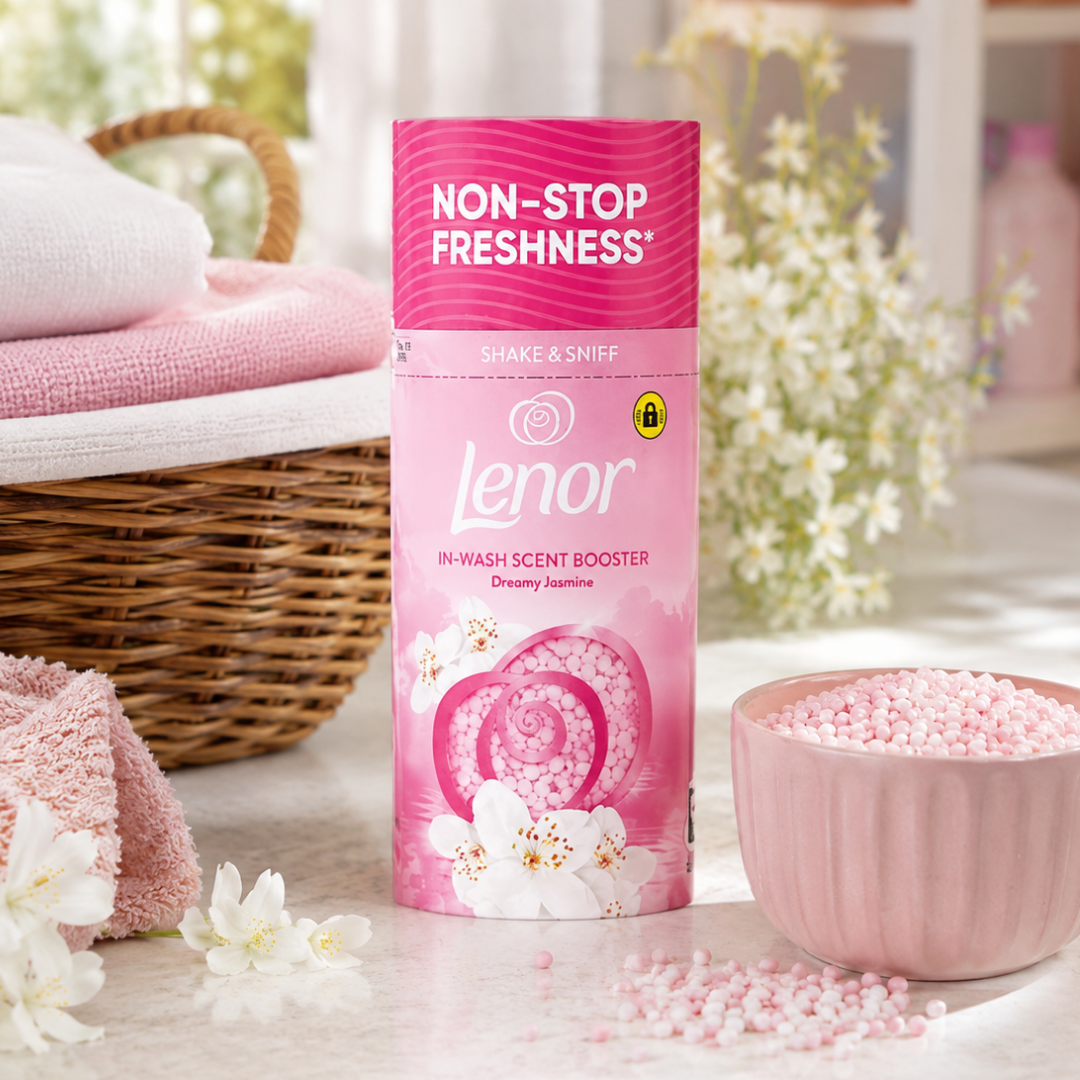 A pink Lenor in-wash scent booster tube stands on a light surface, surrounded by pink fragrance beads in a pink bowl, folded pink towels, and white flowers in a softly lit indoor setting.