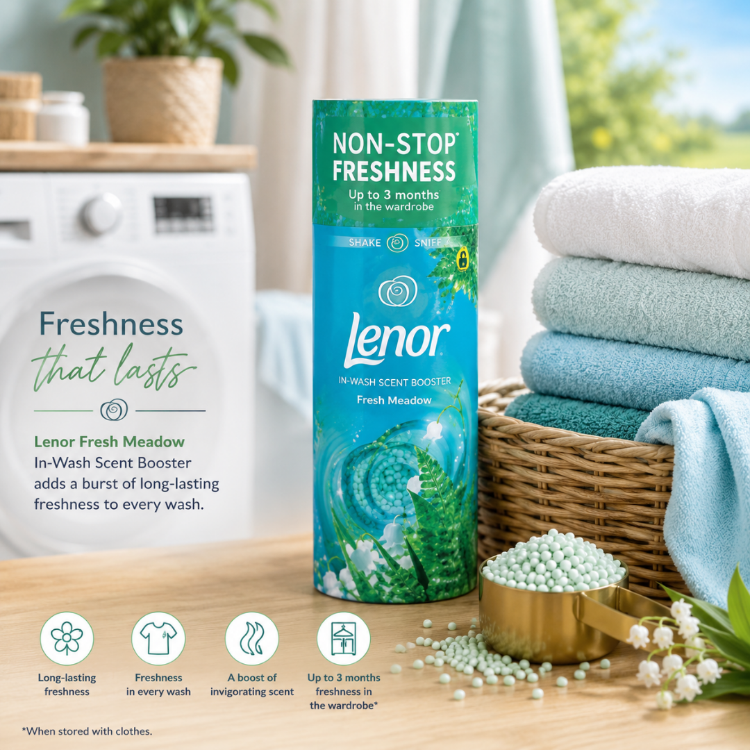 A Lenor Fresh Meadow non-stop freshness in-wash scent booster bottle stands on a wooden surface in a bright laundry room setting, surrounded by folded towels, a basket, a scoop of fragrance beads, and informational text about long-lasting freshness.