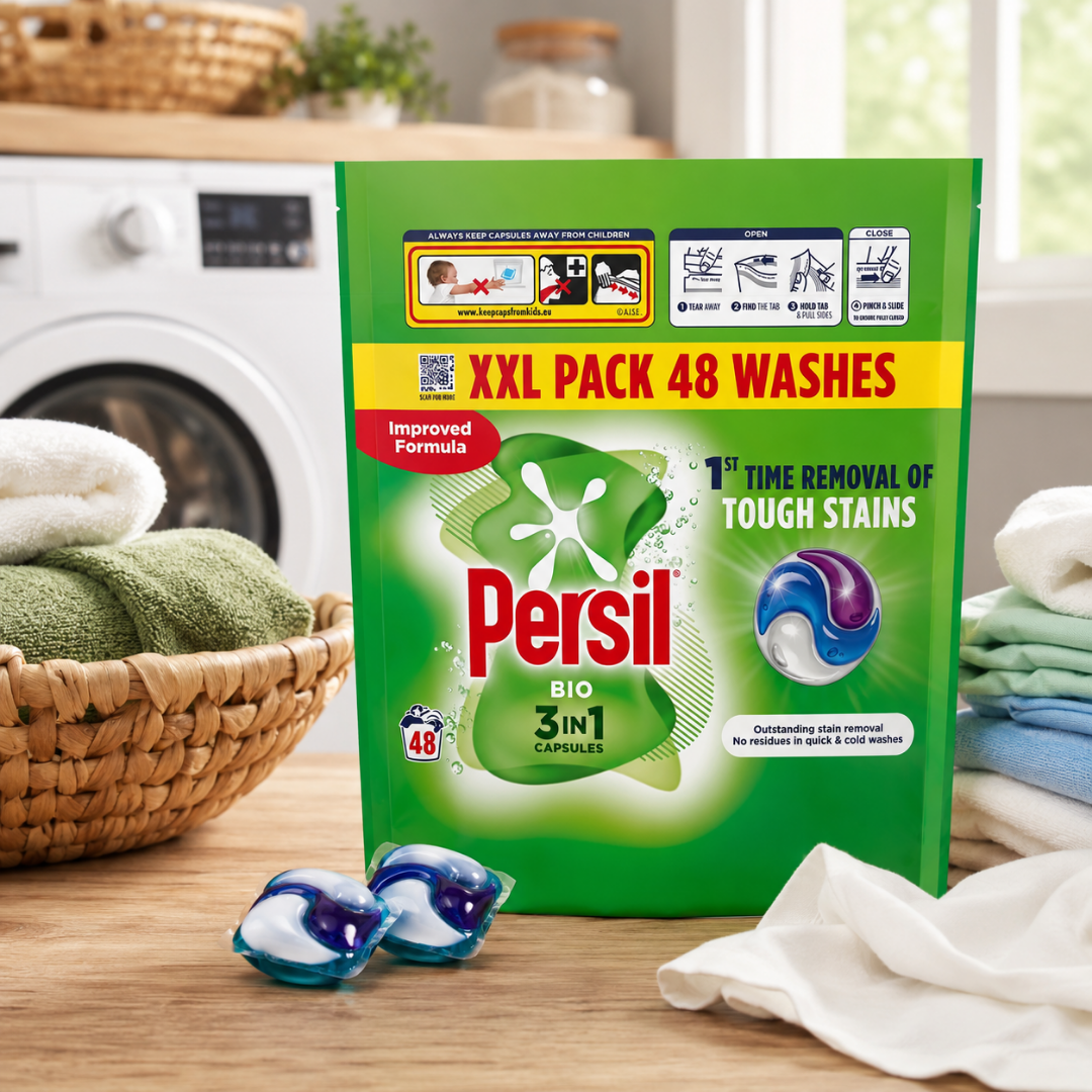 Persil Bio 3-in-1 XXL Pack 48 Washes laundry detergent package standing on a wooden surface, with two blue and white gel capsules in front and folded white and green towels nearby. A washing machine and basket are blurred in the background.