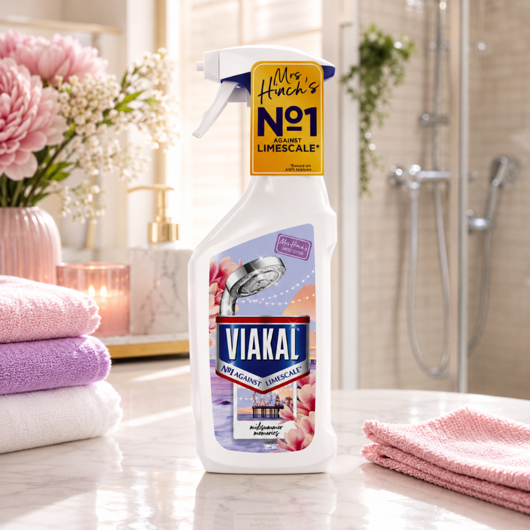 A bottle of VIAKAL shower head cleaner stands on a bathroom counter, with pink flowers, folded towels, and a shower area blurred in the background. The bottle has a white body with a yellow 'Mrs.