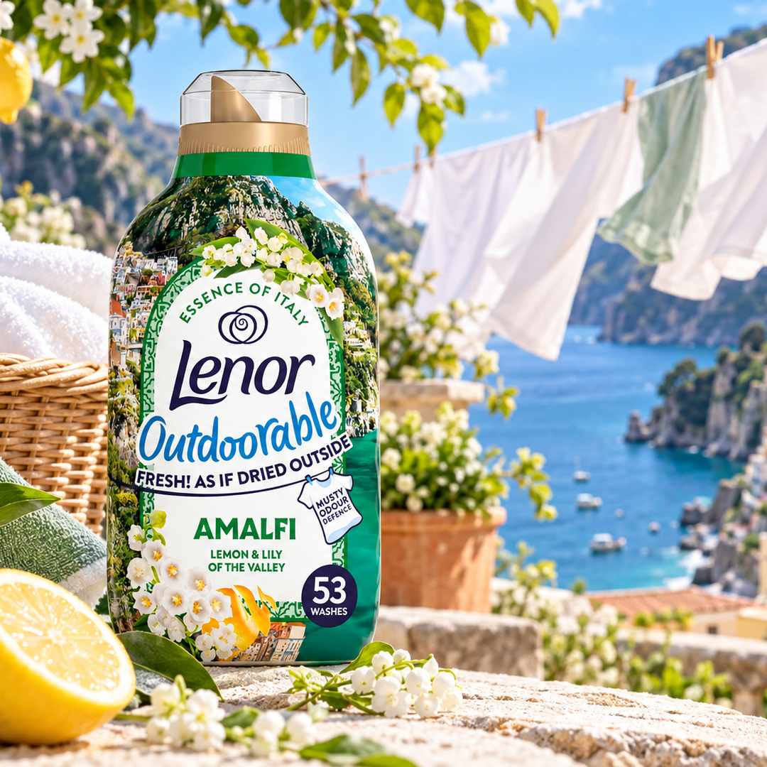 A bottle of Lenor Outdoorable Amalfi laundry detergent stands on a stone ledge with a Mediterranean coastal scene in the background, including blue sea, cliffs, hanging white laundry, flowers, a lemon, and a basket.